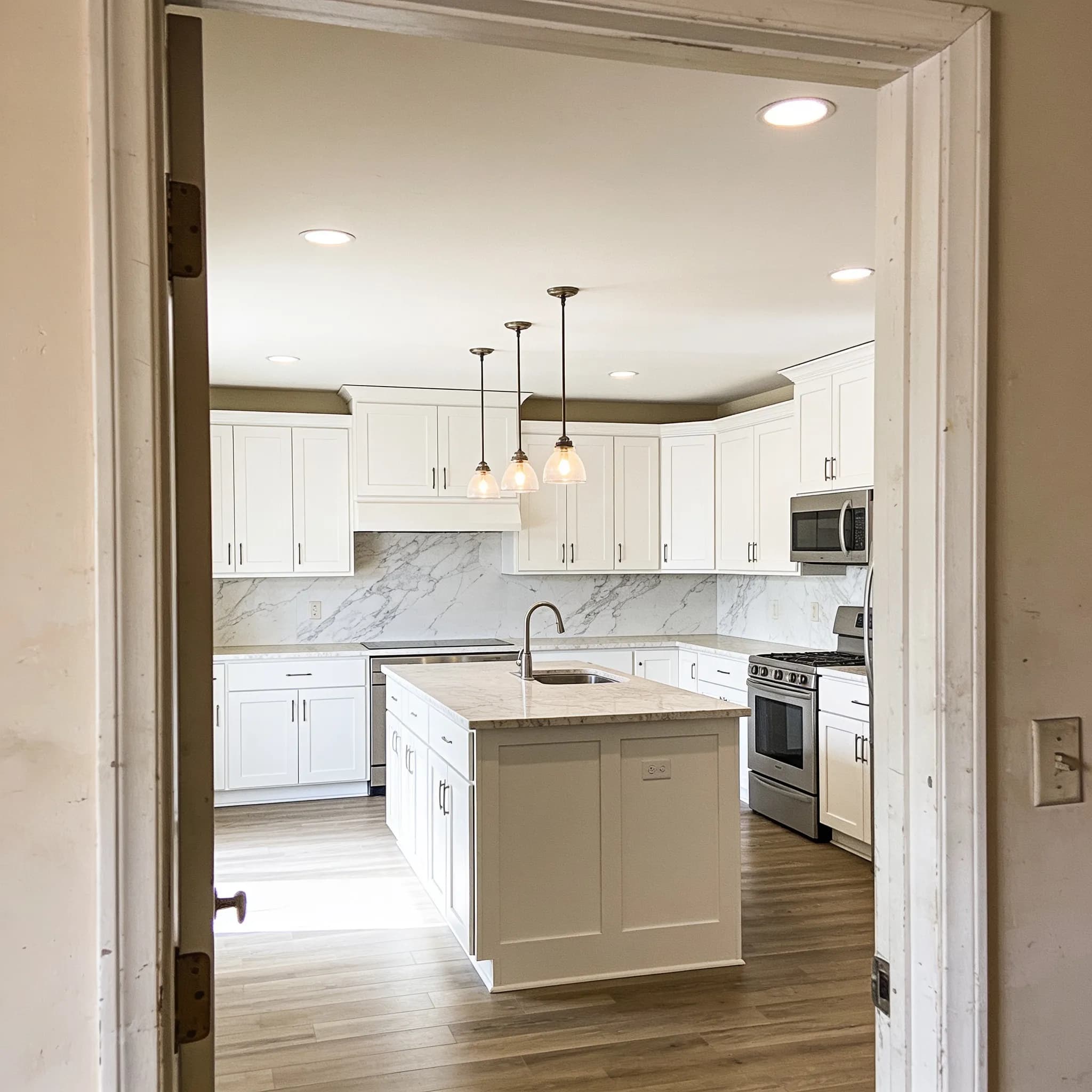 After: The same doorway view, transformed — crisp white shaker cabinets, veined quartz counters, and a new pendant-lit island. Finished on schedule, swept clean every day.